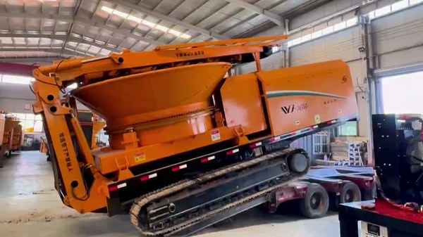 Tracked tub grinder positioned on transport equipment in the workshop; deployment-cost illustration, not transport approval or an included commercial service. Tracked tub grinder positioned on transport equipment in the workshop; deployment-cost illustration, not transport approval or an included commercial service.